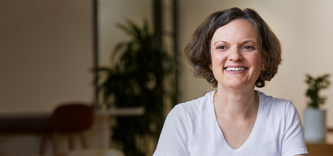 Ruth smiling, sitting indoors with plants in the background.