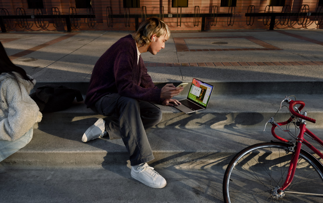 Vysokoškolák sedí na schodech vedle kola a používá iPhone a MacBook