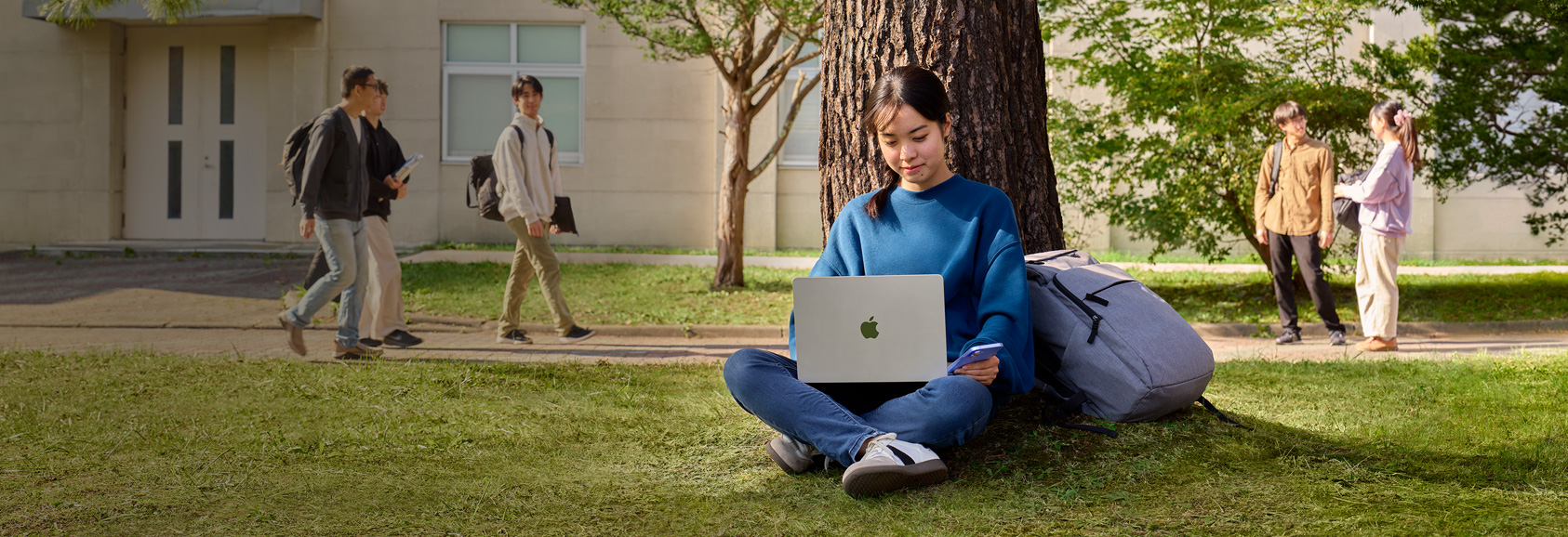 大学のキャンパスで大きな木の前に座っている学生がMacBook AirとiPhoneを使っている。