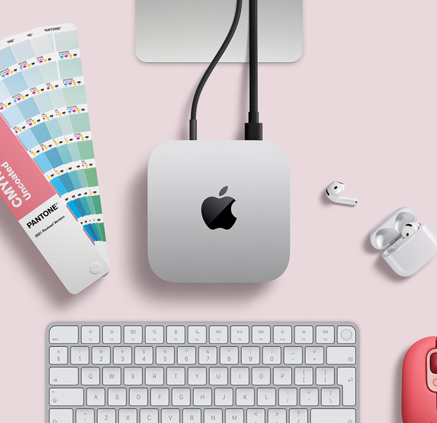 Top view of silver Mac mini in a desk setup with AirPods, a pink wireless keyboard, a pink wireless mouse, and a color swatch fan