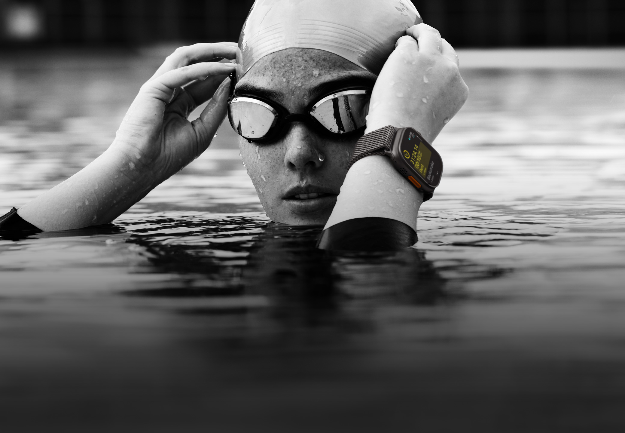 A swimmer’s head and hands above water in a swimming pool wearing Apple Watch Ultra 2 and adjusting goggles.