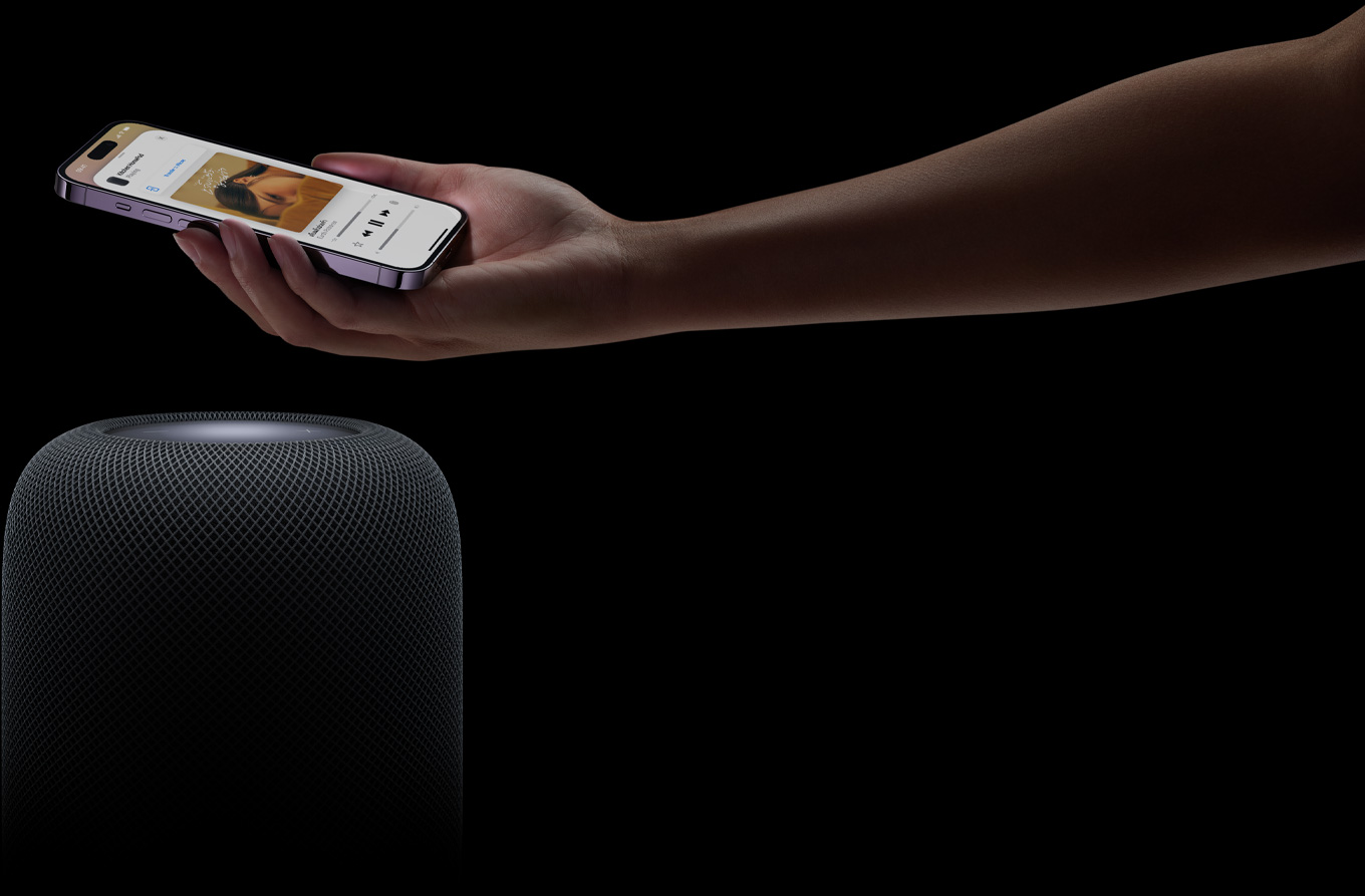 มือข้างหนึ่งยื่นมาจากทางขวาของเว็บเพจโดยที่ในมือถือ iPhone อยู่เหนือลำโพง HomePod