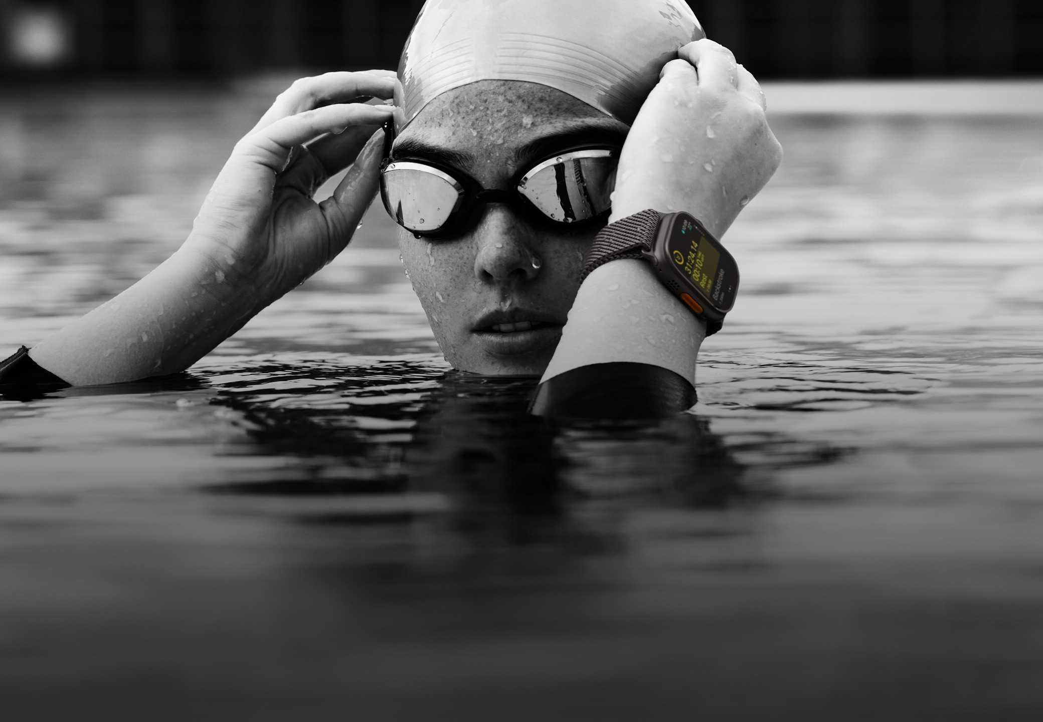 A swimmer’s head and hands above water in a swimming pool wearing Apple Watch Ultra 2 and adjusting goggles.